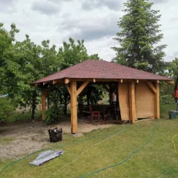 Drewniana altana ogrodowa z czerwoną dachówką bitumiczną, częściowo osłonięta bambusowymi roletami, otoczona zielonym trawnikiem i drzewami, z widocznym w tle domem i rozłożonym parasolem.