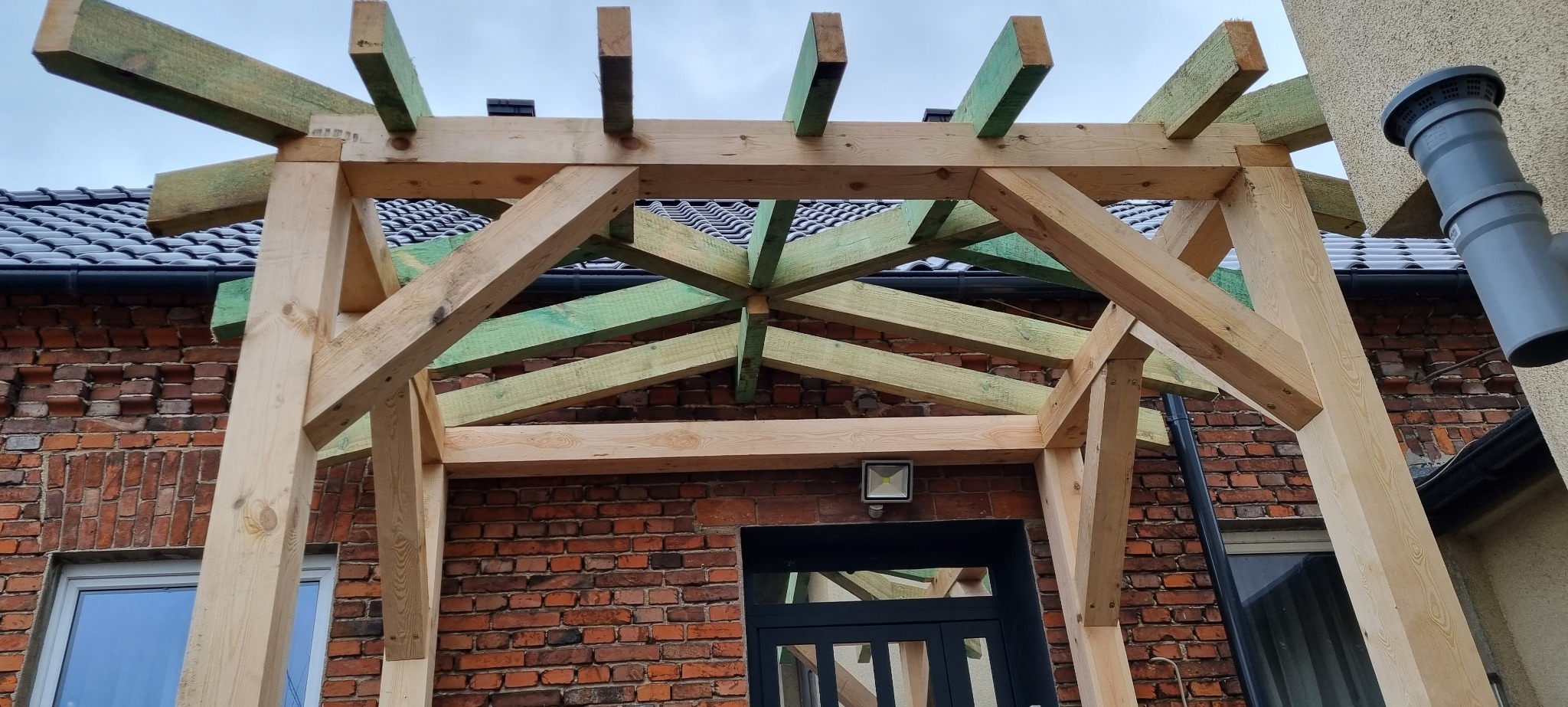 Konstrukcja drewnianej pergoli w trakcie montażu, widoczne impregnowane na zielono elementy, na tle ceglanego budynku z czarną stolarką okienną i dachówką.