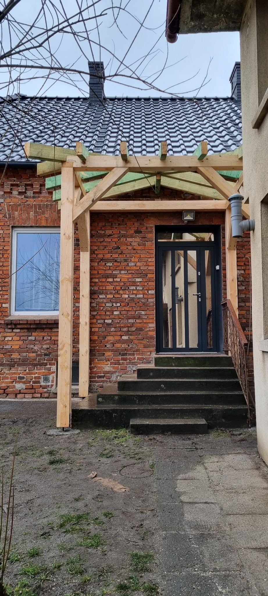 Surowa drewniana konstrukcja pergoli przed wejściem do domu z czerwonej cegły, widoczne betonowe schody i fragment dachu z czarnej dachówki.