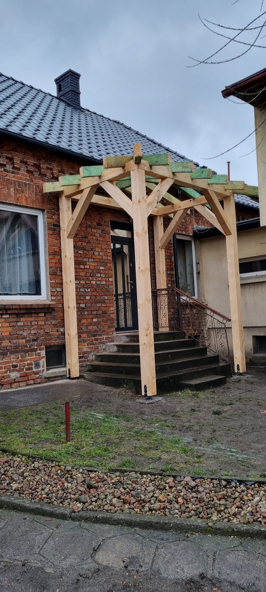 Surowa drewniana konstrukcja z zielonymi elementami montażowymi w trakcie budowy, wzniesiona przed wejściem do domu z czerwonej cegły, widoczne schody wejściowe i fragment posesji z kamieniami.
