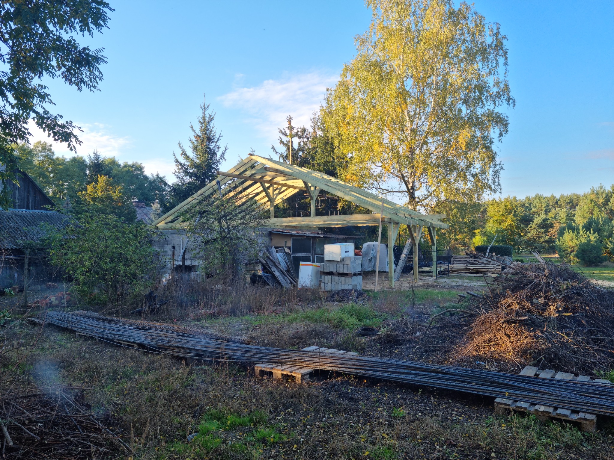 Konstrukcja drewniana z belek na etapie budowy, widok z zewnątrz na tle drzew i niebieskiego nieba, w pobliżu składowane materiały budowlane.