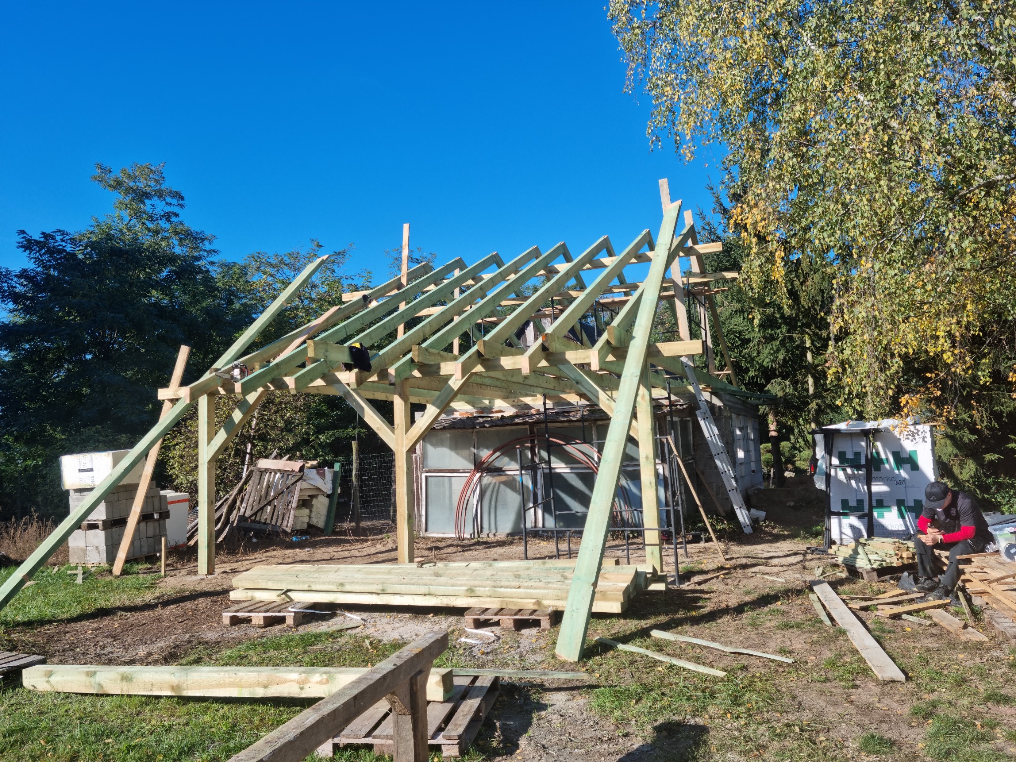 Konstrukcja drewniana w trakcie budowy, widoczne elementy szkieletu dachu i podstawa, w tle budynek gospodarczy i drzewa, na pierwszym planie porozrzucane deski i pracownik w trakcie przerwy.