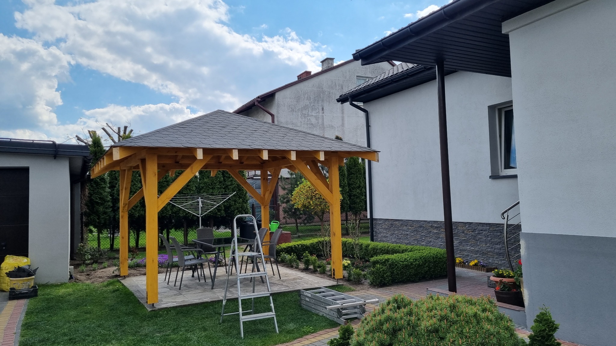 Żółta drewniana konstrukcja z szarym dachem w ogrodzie, widoczne krzesła i stół, drabina oparta o konstrukcję, w tle dom i krzewy.
