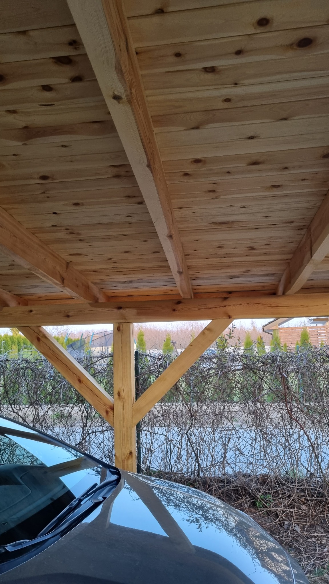 Drewniana konstrukcja zadaszenia nad miejscem parkingowym, widok od dołu, fragment samochodu w kadrze.