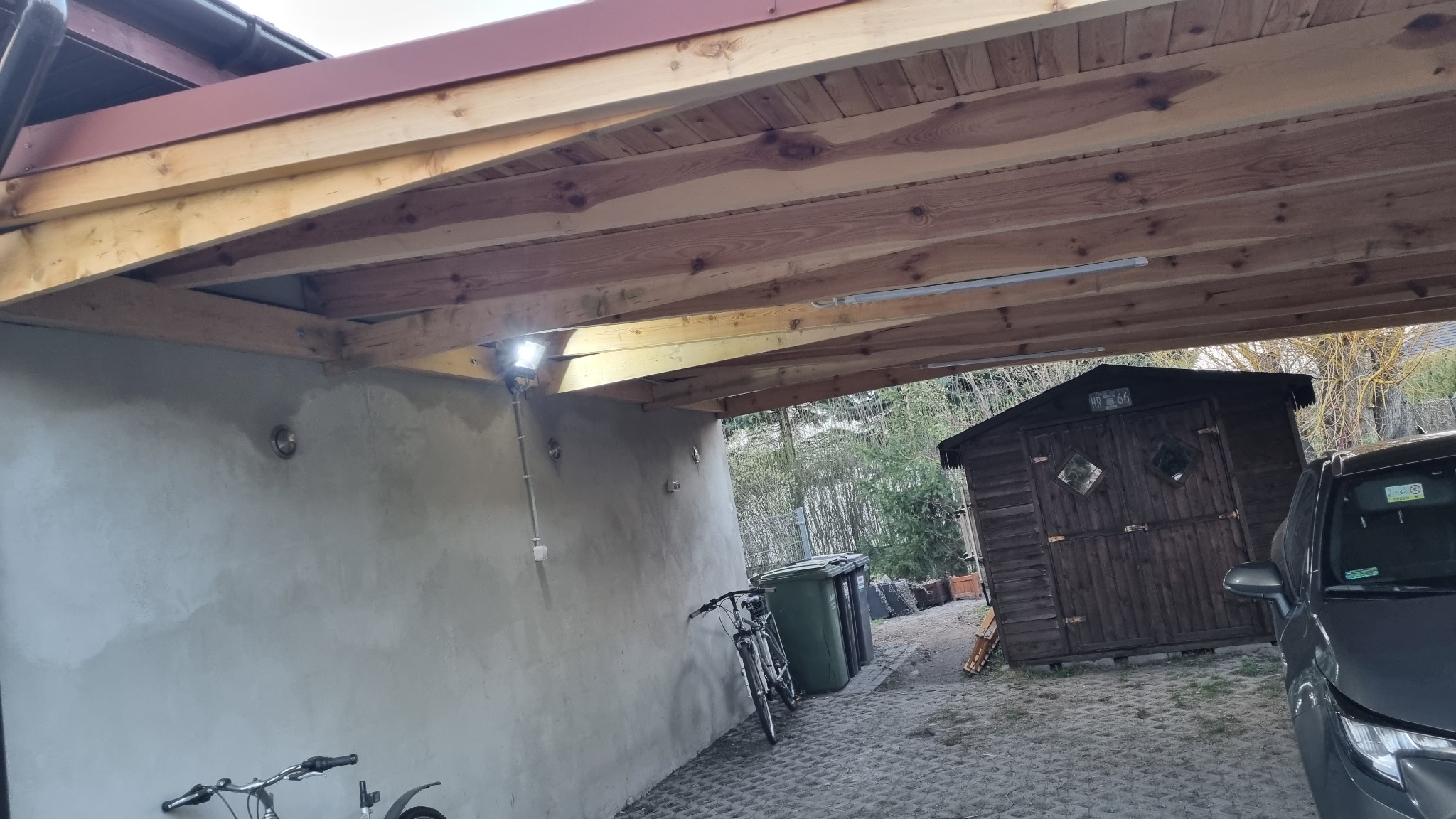 Zadaszenie z drewnianą konstrukcją nośną i oświetleniem LED, widok na podwórze z szopą i samochodem, rower oparty o ścianę.