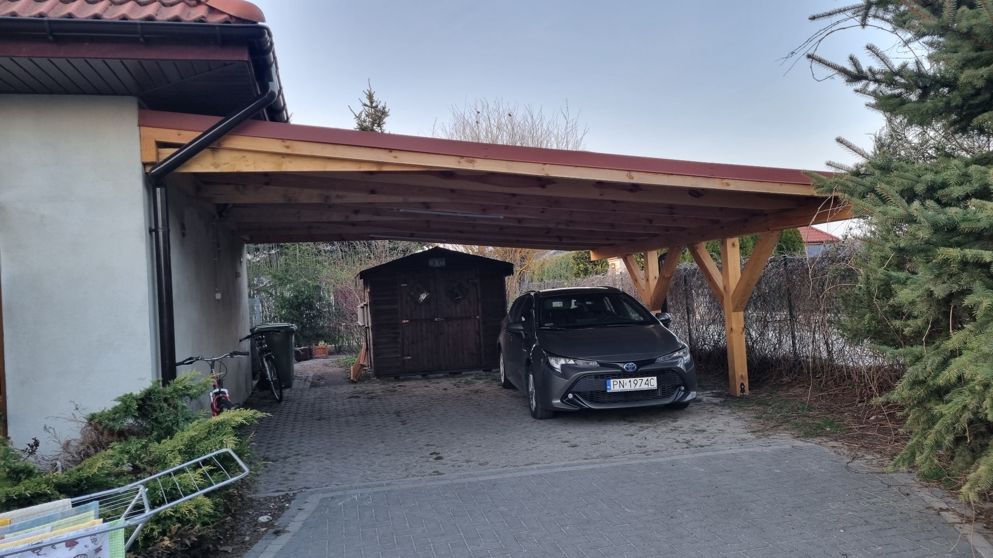 Zadaszenie drewniane w Tuliszkowie, wsparte na solidnych, drewnianych słupach, chroniące zaparkowany samochód osobowy; w tle widoczna drewniana szopa.