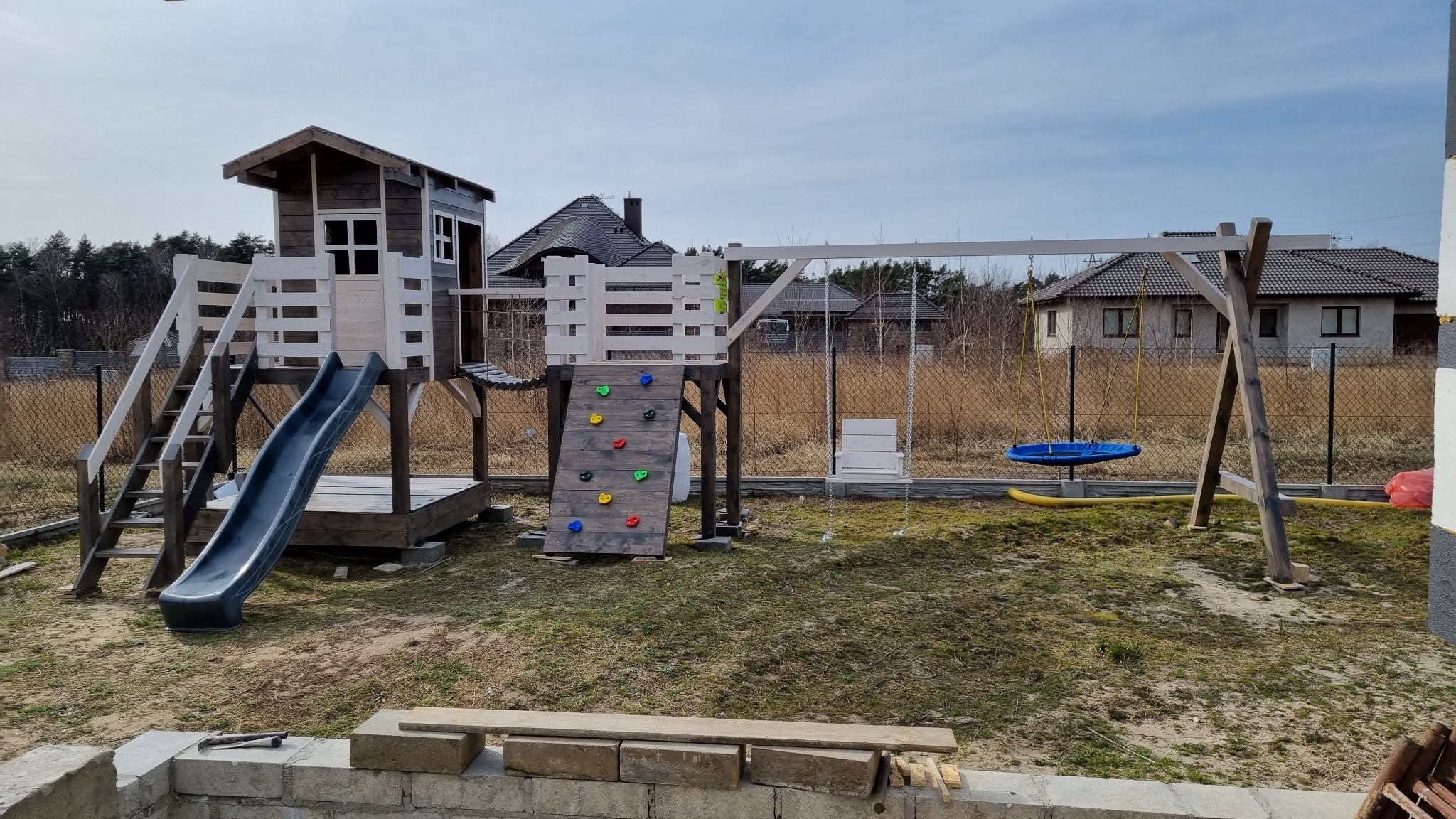 Drewniany plac zabaw z domkiem, zjeżdżalnią, ścianką wspinaczkową i huśtawkami na tle domu jednorodzinnego i ogrodzenia z siatki.