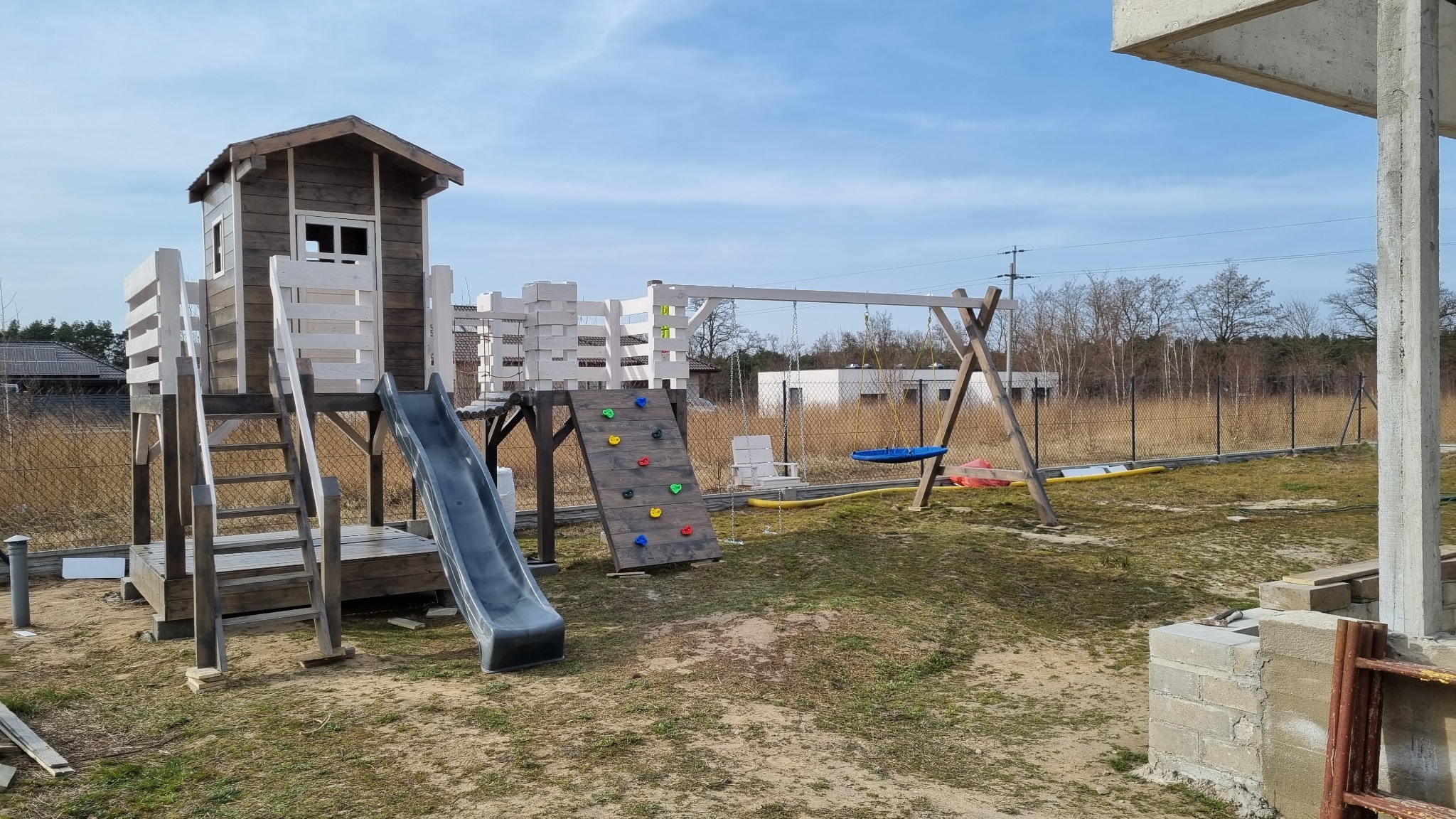 Drewniany plac zabaw z domkiem, zjeżdżalnią, ścianką wspinaczkową i huśtawką na trawiastym podwórku, częściowo w cieniu niedokończonej konstrukcji.