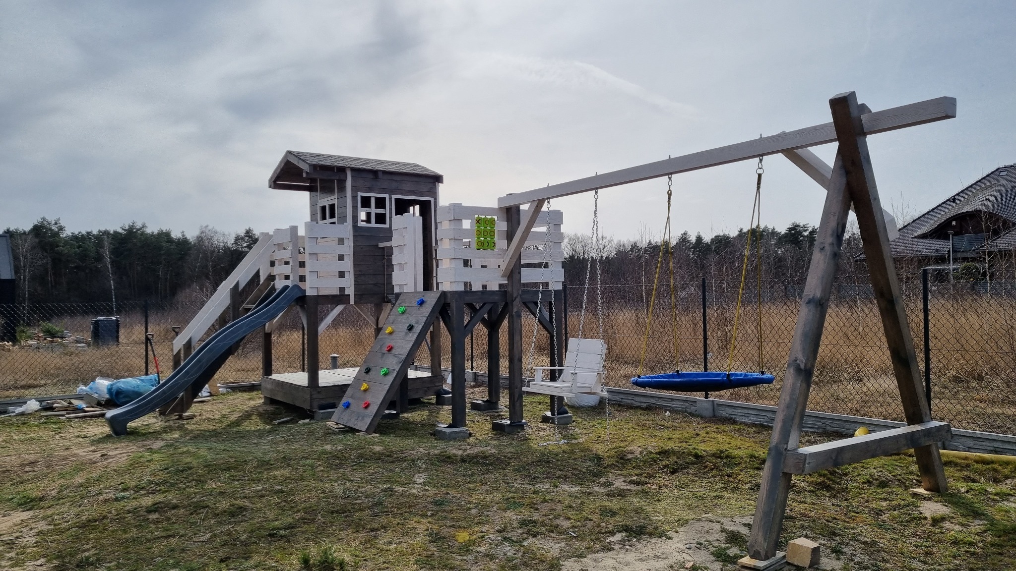 Drewniany plac zabaw z domkiem, zjeżdżalnią, ścianką wspinaczkową i huśtawkami na tle zalesionego ogrodu, częściowo na podmurówce z betonowych elementów.