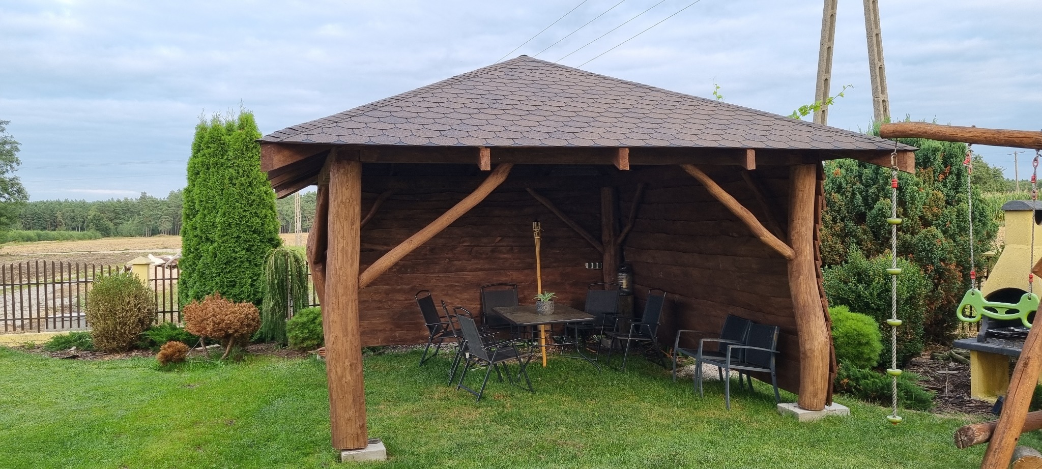 Drewniana pergola z brązowym dachem z gontów bitumicznych, wyposażona w stół i czarne krzesła na trawniku; w tle widoczne drzewa i huśtawka.