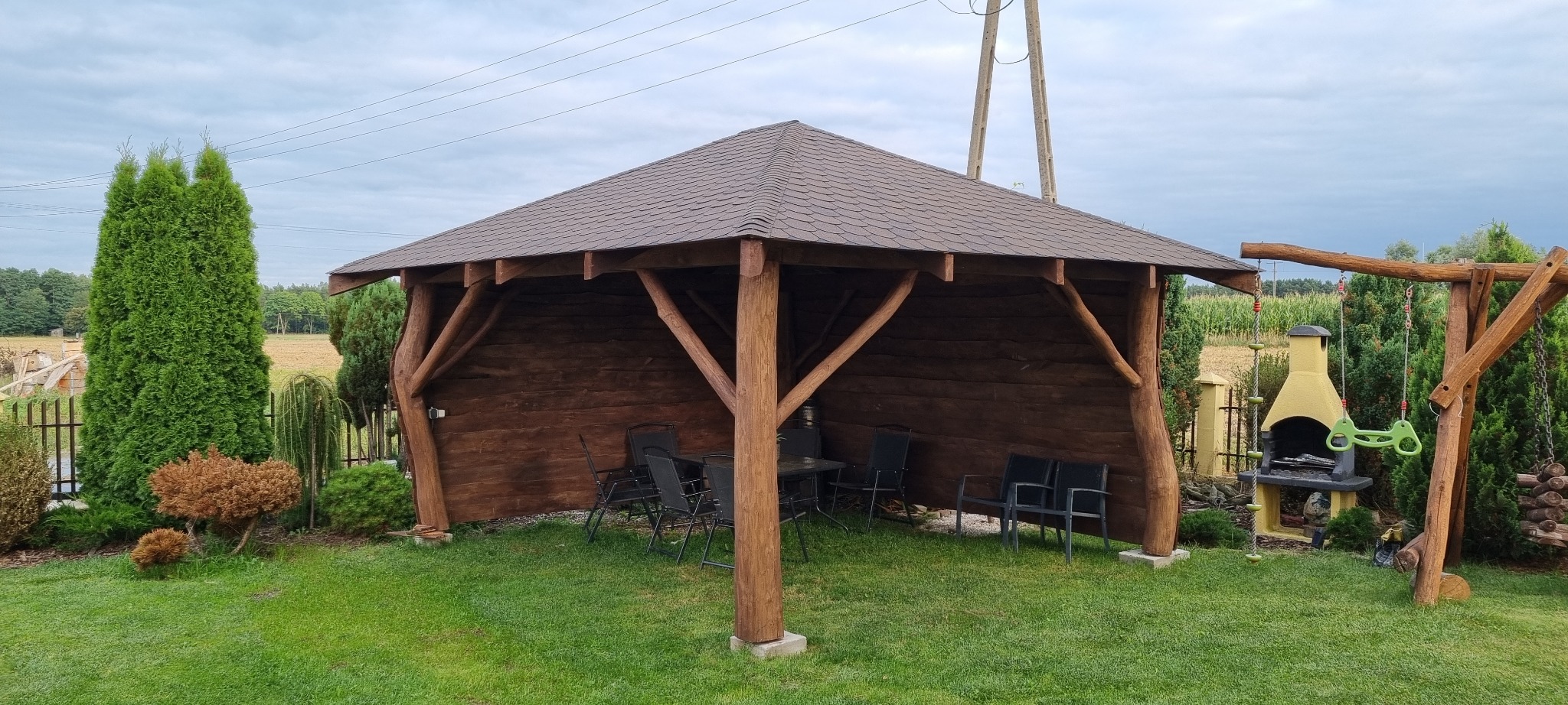 Drewniana altana z brązowym dachem krytym gontem bitumicznym, wyposażona w czarne krzesła i stolik, na trawniku, z grillem i huśtawką w tle.