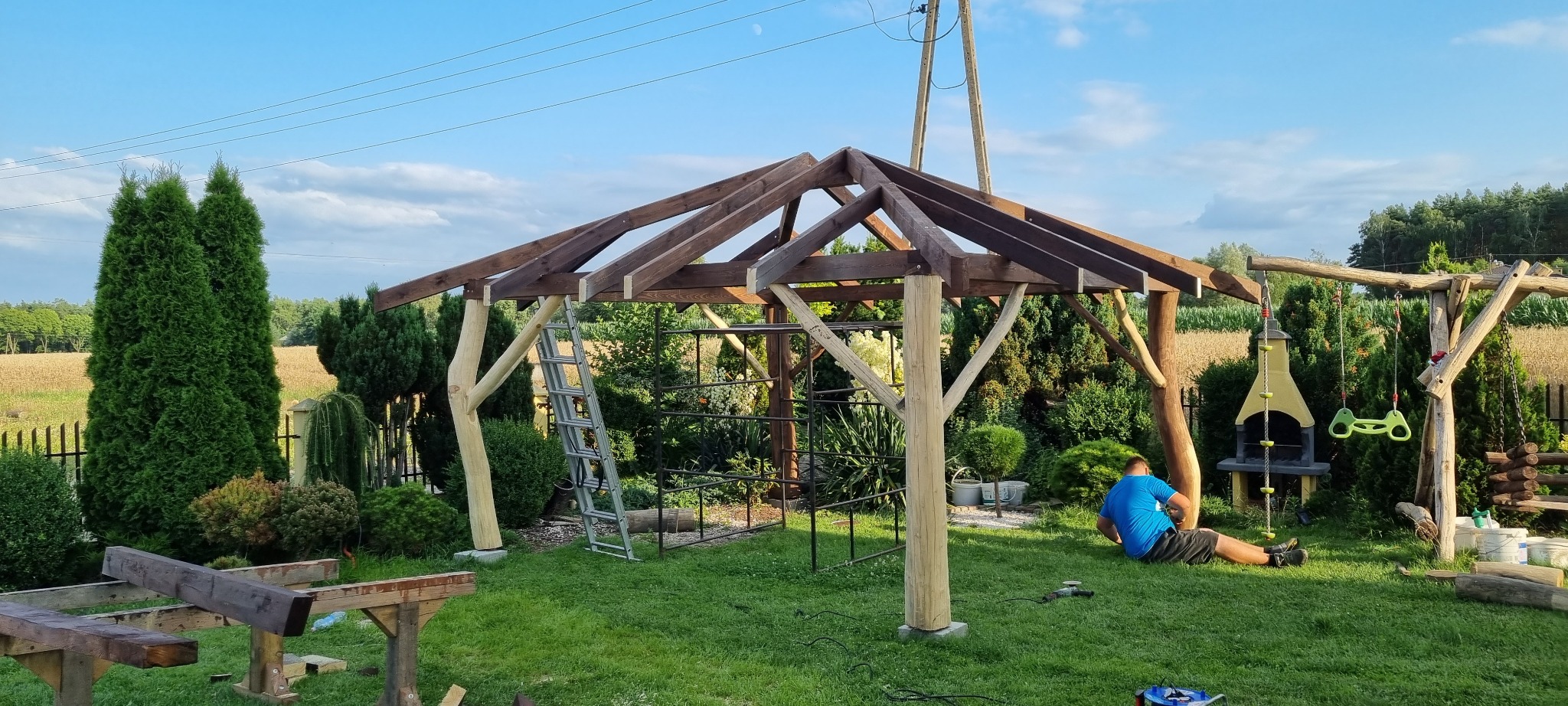 Konstrukcja drewnianej pergoli w trakcie montażu na trawniku, widoczna drabina oparta o konstrukcję i mężczyzna w niebieskiej koszulce siedzący na trawie obok słupa pergoli.