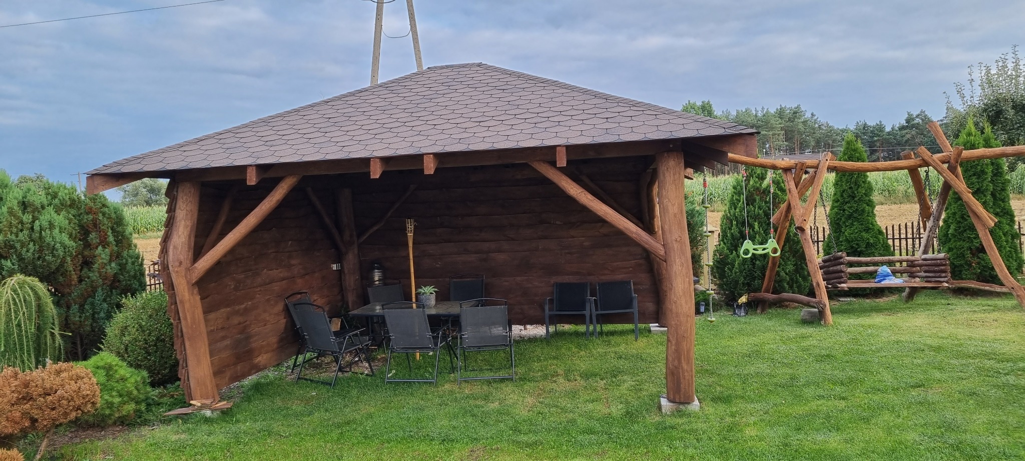 Drewniana altana z ciemnobrązowym dachem z gontów bitumicznych, wyposażona w stół i krzesła, na trawniku w ogrodzie, obok huśtawka z bali.