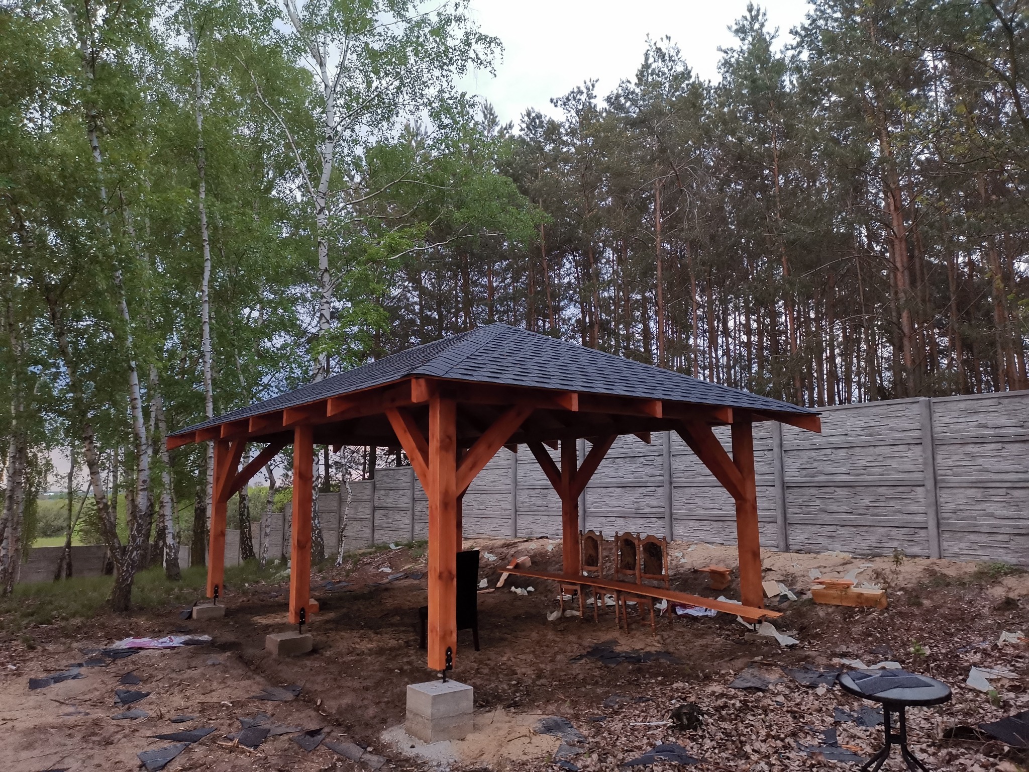 Nowo postawiona drewniana konstrukcja z ciemnym, spadzistym dachem z gontu bitumicznego, wsparta na betonowych fundamentach, otoczona drzewami i betonowym ogrodzeniem.