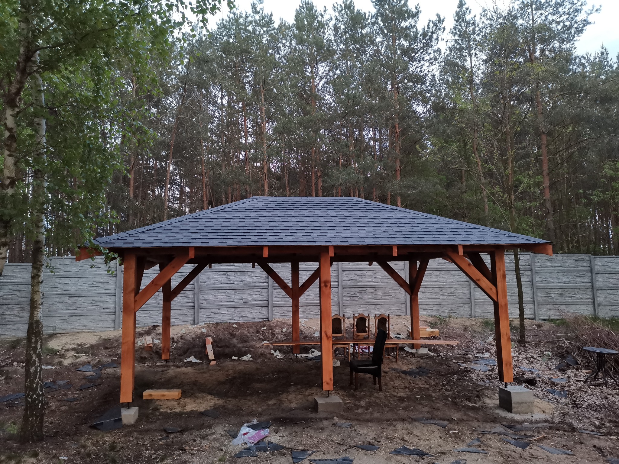 Drewniana pergola z ciemnym dachem bitumicznym na tle lasu i betonowego ogrodzenia, częściowo umeblowana, w trakcie prac wykończeniowych.