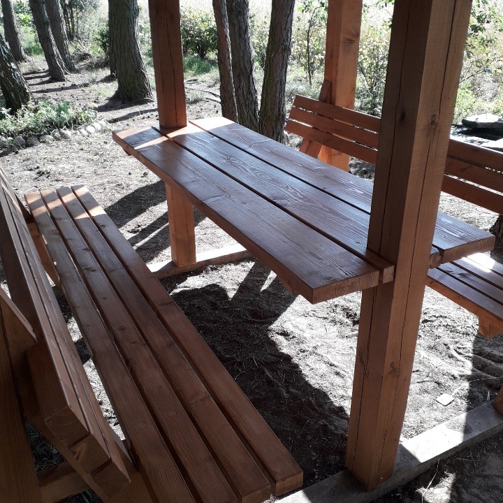 Drewniana konstrukcja z ławkami i stołem na zewnątrz, rzucająca cienie na ziemię, otoczona drzewami.