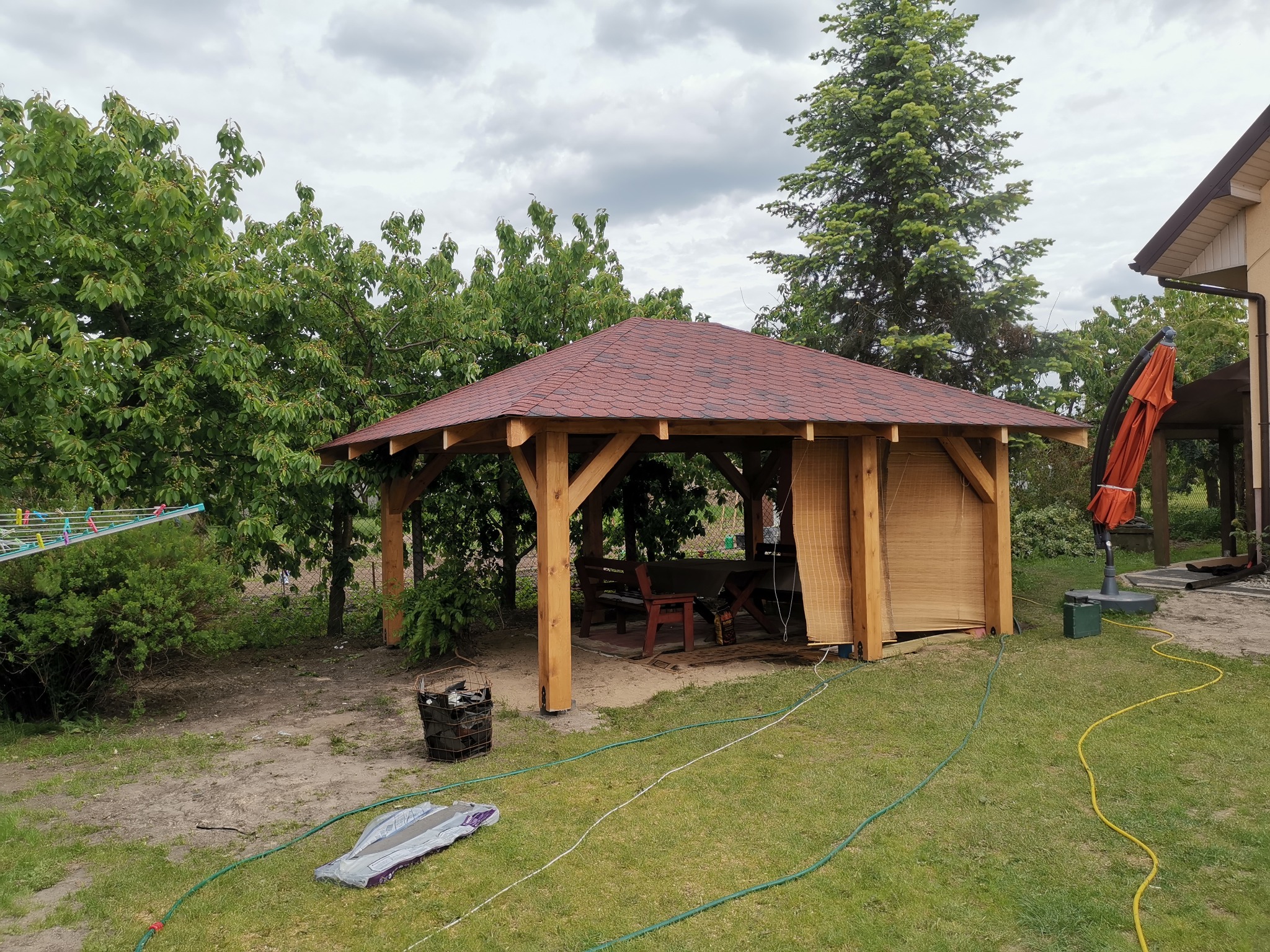 Drewniana altana ogrodowa z czerwoną dachówką bitumiczną, częściowo osłonięta bambusowymi roletami, otoczona zielonym trawnikiem i drzewami, z widocznym w tle domem i rozłożonym parasolem.