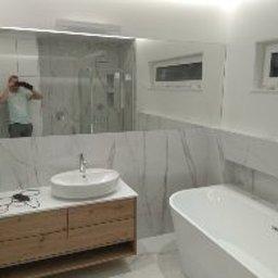 Adam Woźniak Usługi Remontowo Budowlane - Nowoczesna łazienka z dużym lustrem, białą umywalką nablatową na drewnianej szafce, białą wanną i marmurowymi płytkami. W lustrze odbija się osoba robiąca zdjęcie.