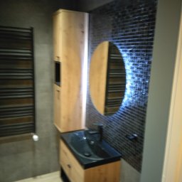 Adam Woźniak Usługi Remontowo Budowlane - Nowoczesna łazienka z drewnianą szafką pod umywalkę z czarnym blatem, okrągłym lustrem z podświetleniem LED i grzejnikiem drabinkowym na szarej ścianie.