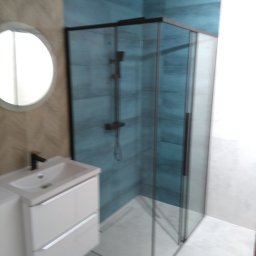 Adam Woźniak Usługi Remontowo Budowlane - Nowoczesna łazienka z kabiną prysznicową typu walk-in z czarnymi profilami, białą umywalką z szafką i okrągłym lustrem na ścianie wykończonej w beżowe wzory. Ściany kabiny wyłożone niebieskimi...