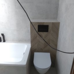 Adam Woźniak Usługi Remontowo Budowlane - Biała wanna i toaleta podwieszana z czarnym przyciskiem spłukującym na tle płytek imitujących drewno ułożonych w jodełkę, w jasnej łazience wykończonej szarymi płytkami.