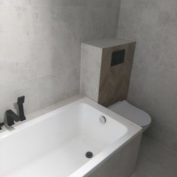 Adam Woźniak Usługi Remontowo Budowlane - Nowoczesna łazienka z białą wanną, czarną armaturą i toaletą w zabudowie z drewnopodobną mozaiką ułożoną w jodełkę na tle szarych płytek.