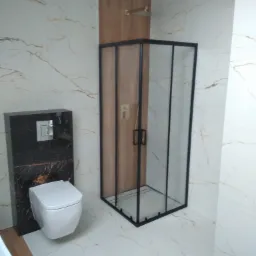 Nowoczesna łazienka z kabiną prysznicową z czarną ramą, podwieszaną toaletą w obudowie z czarnego marmuru i płytkami imitującymi marmur z delikatnym, złotym żyłowaniem na ścianach i podłodze.