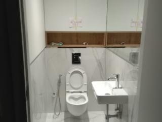 Biała toaleta podwieszana z otwartą klapą, bidetka i umywalka nablatowa na metalowym stelażu w łazience wykończonej białymi płytkami imitującymi marmur. Nad toaletą wnęka z drewnianymi półkami...