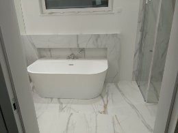 Nowoczesna, biała łazienka z wolnostojącą wanną i kabiną prysznicową, wykończona płytkami imitującymi marmur z delikatnym, złotym żyłkowaniem, widok zza uchylonych drzwi.