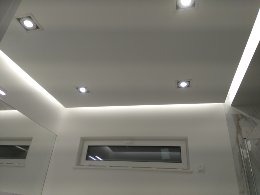 Białe wnętrze łazienki z oświetleniem LED podsufitowym i punktowymi lampami, widoczne okno z białymi ramami oraz fragment lustra po lewej stronie.