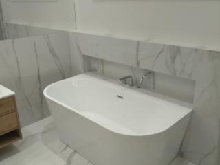 Nowoczesna, wolnostojąca wanna w białej łazience wykończonej płytkami imitującymi marmur z szarymi żyłami. Bateria umieszczona w specjalnej wnęce nad wanną.