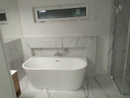 Nowoczesna, biała wanna wolnostojąca z chromowaną armaturą, obudowana płytkami imitującymi marmur, w jasnej łazience z oknem i kabiną prysznicową w tle.