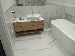 Nowoczesna łazienka z umywalką nablatową na drewnianej szafce, białą wanną wolnostojącą i marmurowymi płytkami na ścianach i podłodze. Widoczne niedokończone podłączenie elektryczne do oświetlenia...