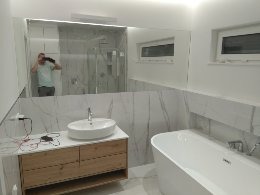 Nowoczesna łazienka z dużym lustrem, białą umywalką nablatową na drewnianej szafce, białą wanną i marmurowymi płytkami. W lustrze odbija się osoba robiąca zdjęcie.