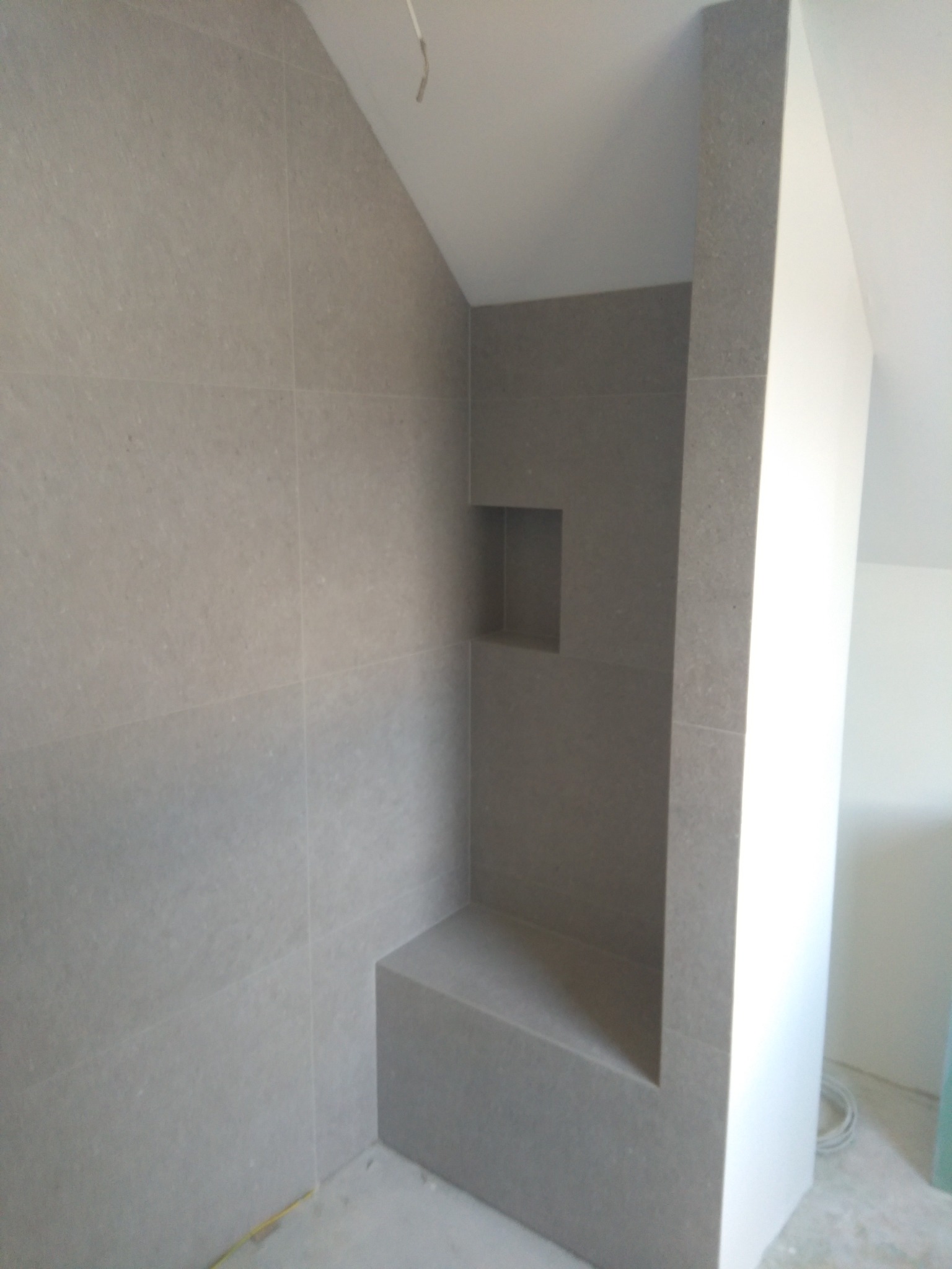 Wykończona wnęka prysznicowa z siedziskiem, wyłożona szarymi płytkami w formacie prostokątnym, z widoczną półką w ścianie i fragmentem sufitu skośnego.