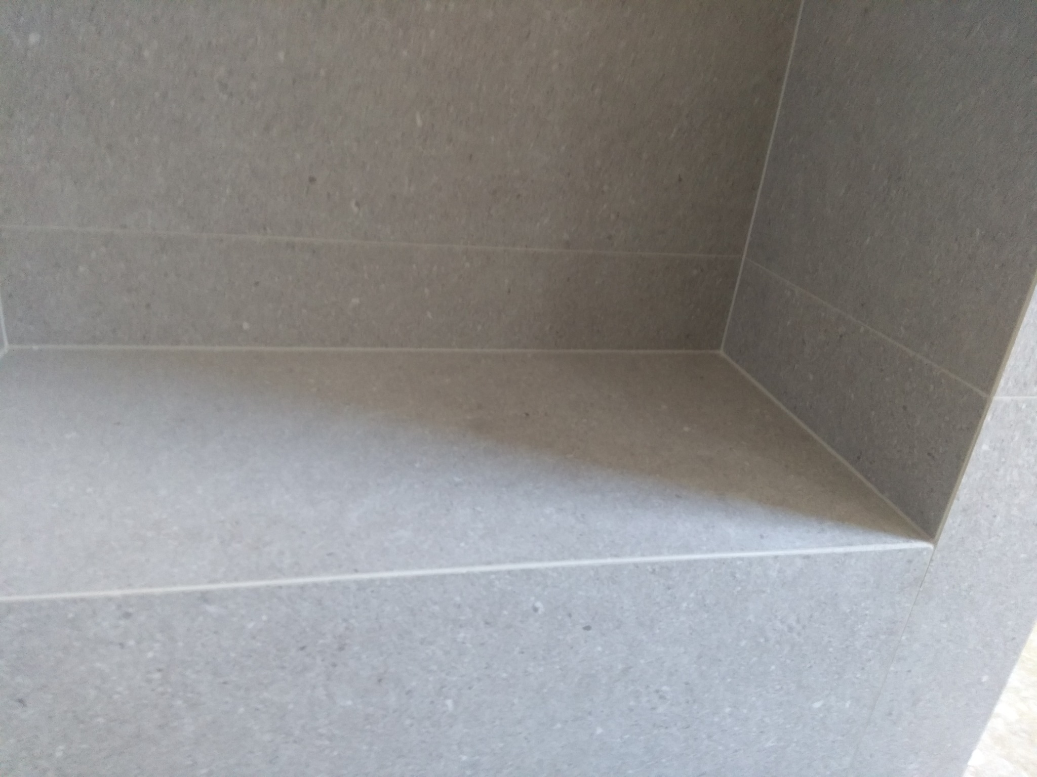 Wnęka w ścianie wyłożona szarymi, matowymi płytkami ceramicznymi z delikatną fugą. Widoczne oświetlenie padające z góry, tworzące cień na dolnej półce wnęki.