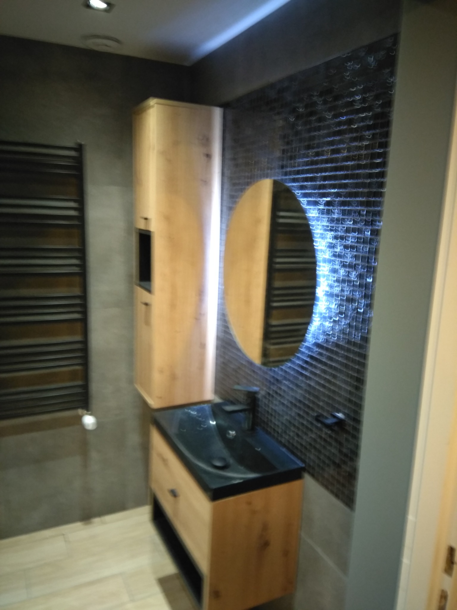 Nowoczesna łazienka z drewnianą szafką pod umywalkę z czarnym blatem, okrągłym lustrem z podświetleniem LED i grzejnikiem drabinkowym na szarej ścianie.