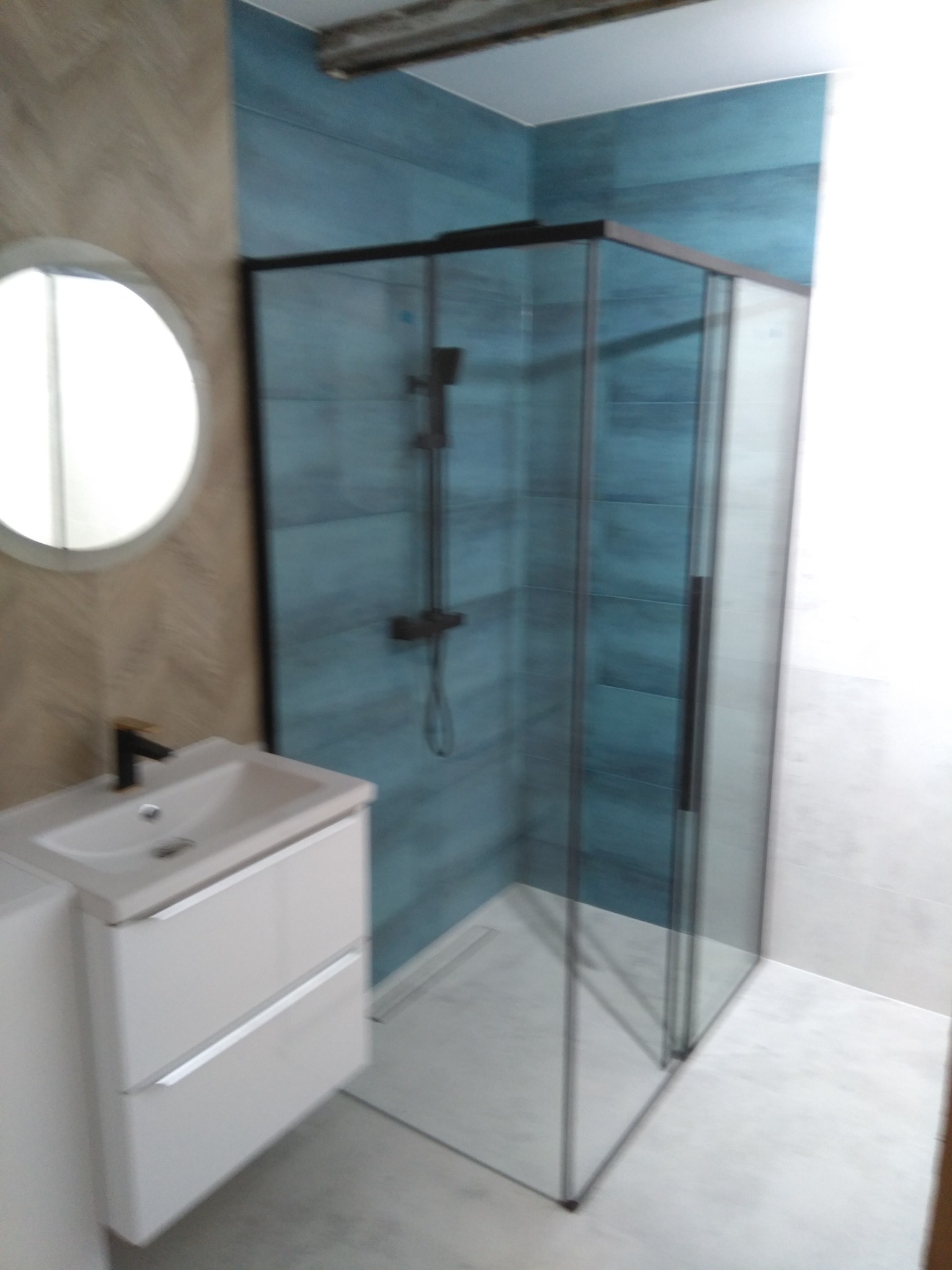 Nowoczesna łazienka z kabiną prysznicową typu walk-in z czarnymi profilami, białą umywalką z szafką i okrągłym lustrem na ścianie wykończonej w beżowe wzory. Ściany kabiny wyłożone niebieskimi...