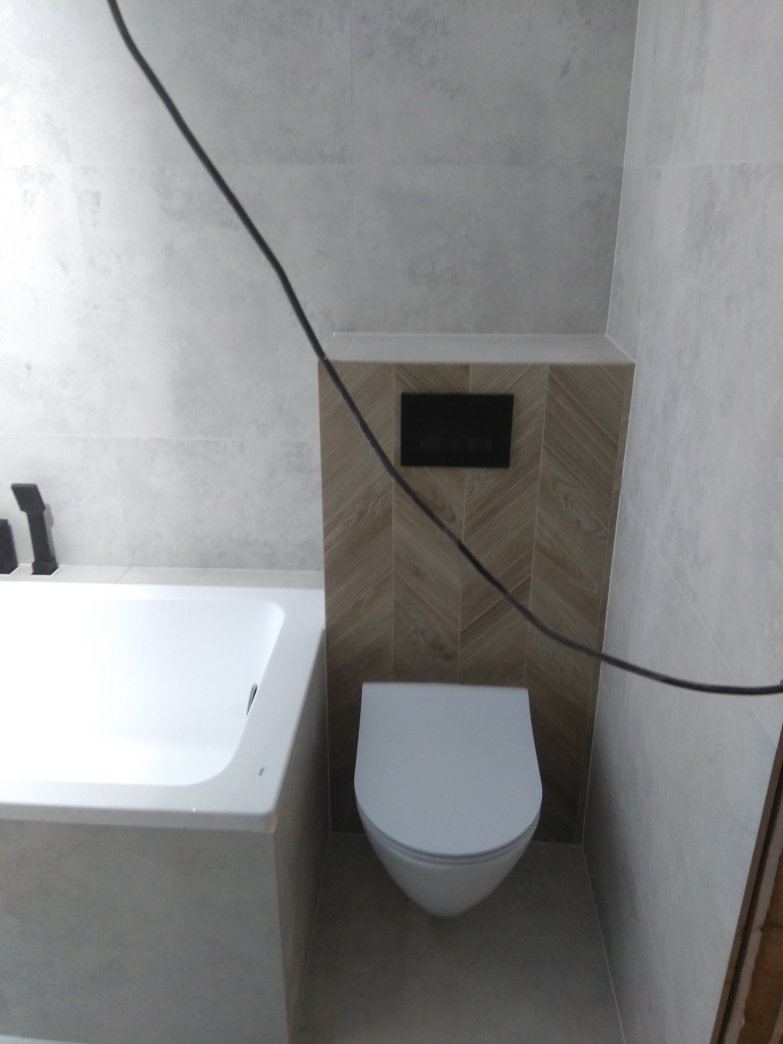 Biała wanna i toaleta podwieszana z czarnym przyciskiem spłukującym na tle płytek imitujących drewno ułożonych w jodełkę, w jasnej łazience wykończonej szarymi płytkami.