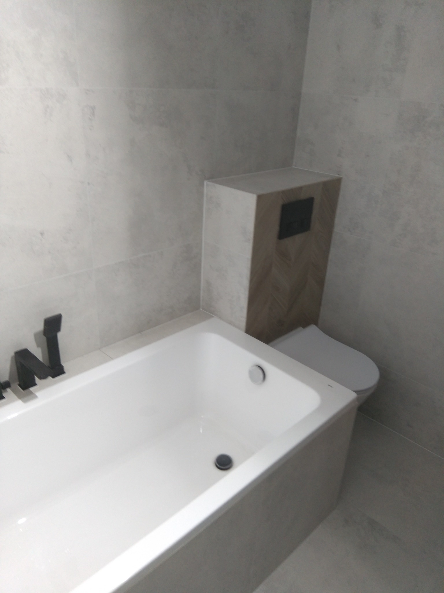 Nowoczesna łazienka z białą wanną, czarną armaturą i toaletą w zabudowie z drewnopodobną mozaiką ułożoną w jodełkę na tle szarych płytek.