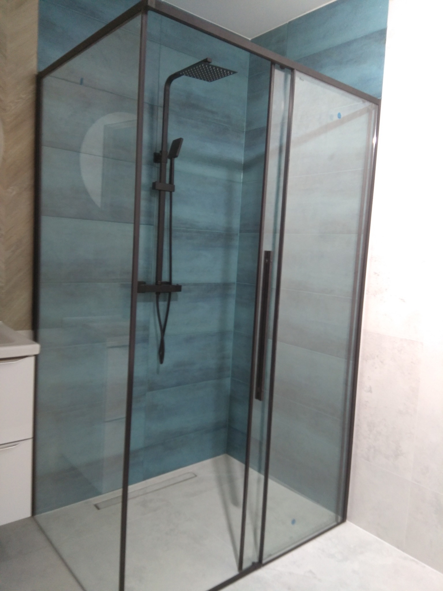 Nowoczesna kabina prysznicowa z czarnymi profilami na tle płytek w odcieniach błękitu i szarości, widoczny odpływ liniowy w posadzce.