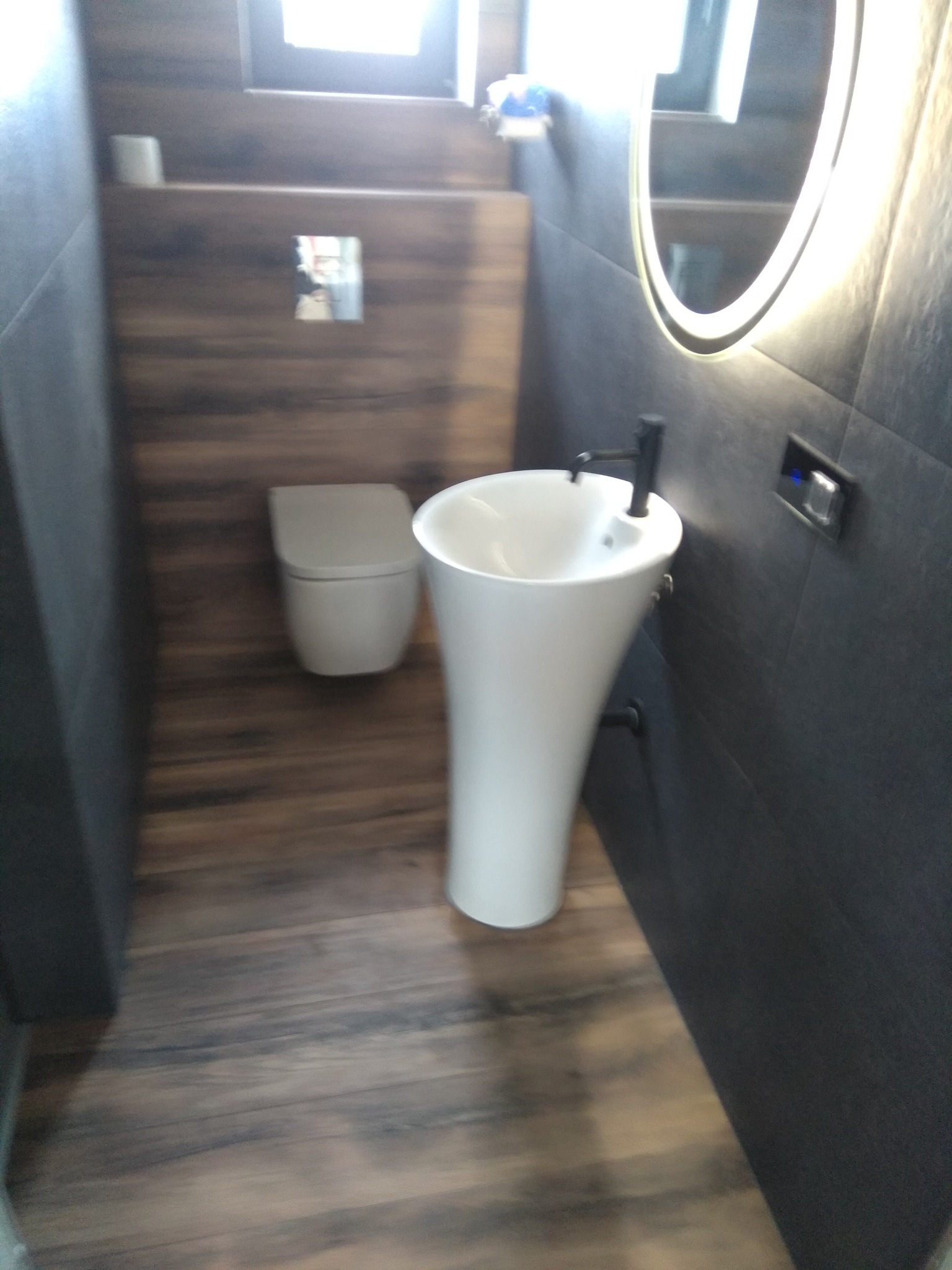 Nowoczesna łazienka z podwieszaną toaletą, umywalką na nodze i podświetlanym lustrem owalnym, wykończona płytkami imitującymi drewno i ciemnymi płytkami.