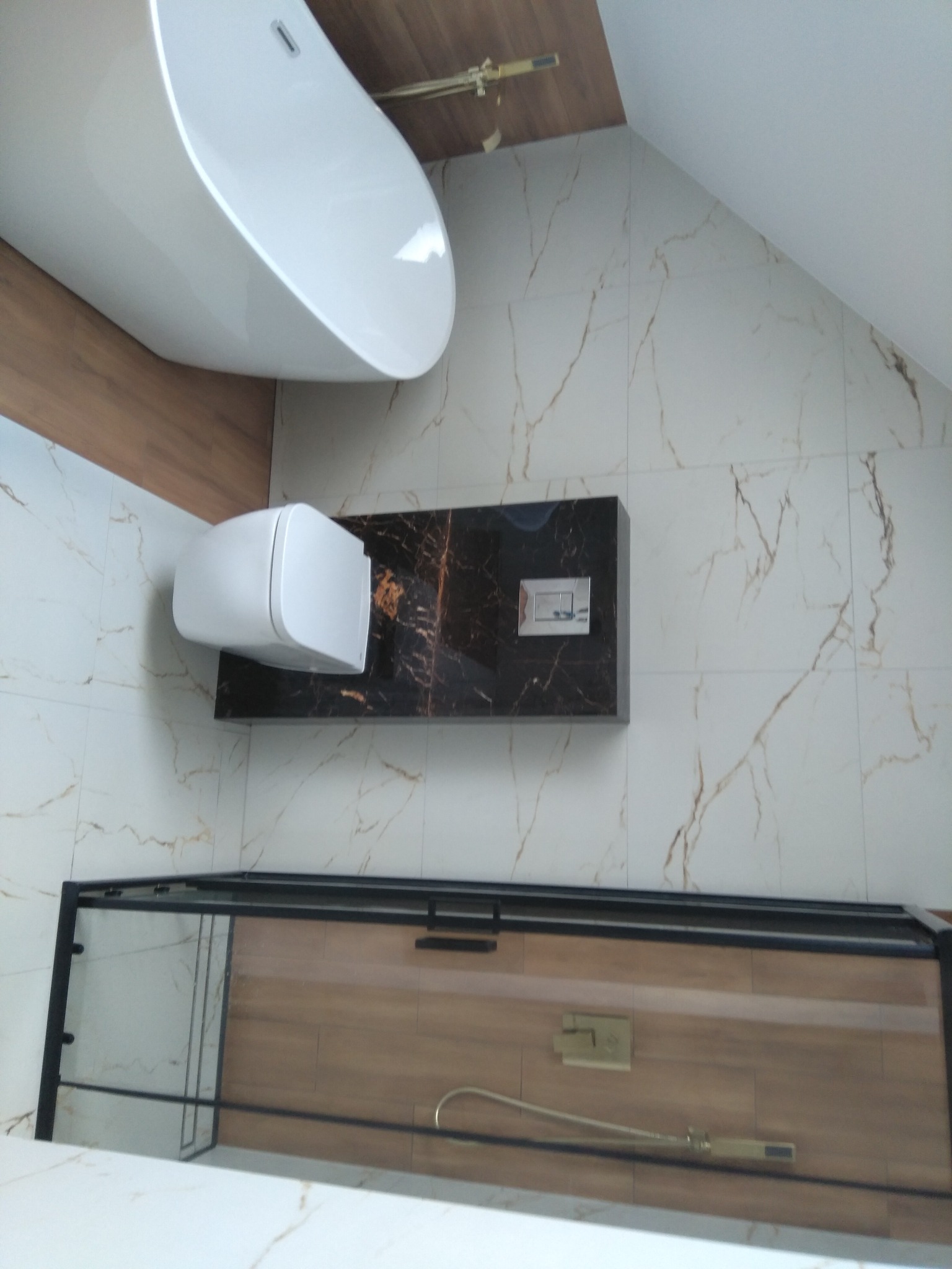 Nowoczesna łazienka w odcieniach bieli i brązu, z wolnostojącą wanną, podwieszaną toaletą na ciemnym blacie i kabiną prysznicową z czarnymi profilami.