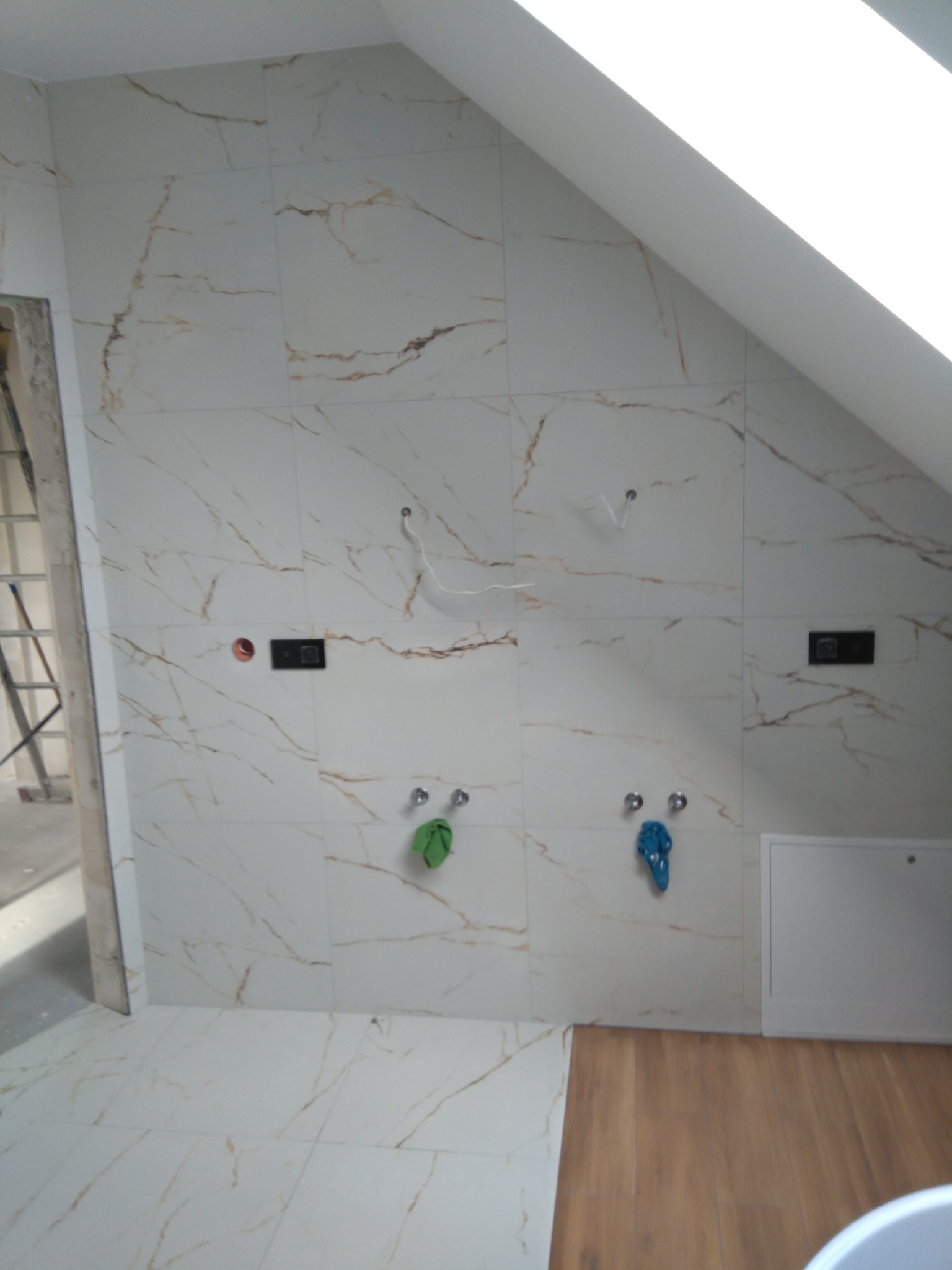 Łazienka w trakcie remontu: ściany i podłoga wyłożone białymi płytkami imitującymi marmur, widoczne wyprowadzenia instalacji wodnej i elektrycznej, fragment drewnianej podłogi oraz drabina malarska.