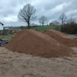 Dwie pryzmy brązowego materiału na ziemi, prawdopodobnie piasku lub ziemi, z widocznymi śladami opon, w tle dom z białymi ścianami i szarym dachem oraz drzewa bez liści pod pochmurnym niebem.