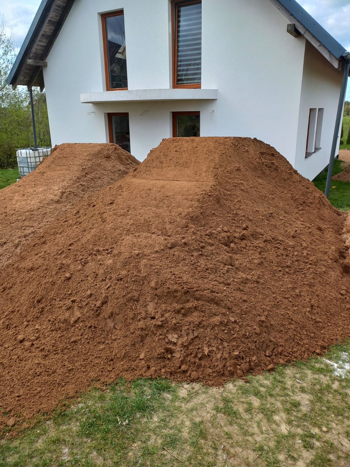 Dwie pryzmy brązowej ziemi usypane przed nowo wybudowanym, białym domem z brązowymi ramami okiennymi, widoczny fragment trawnika.