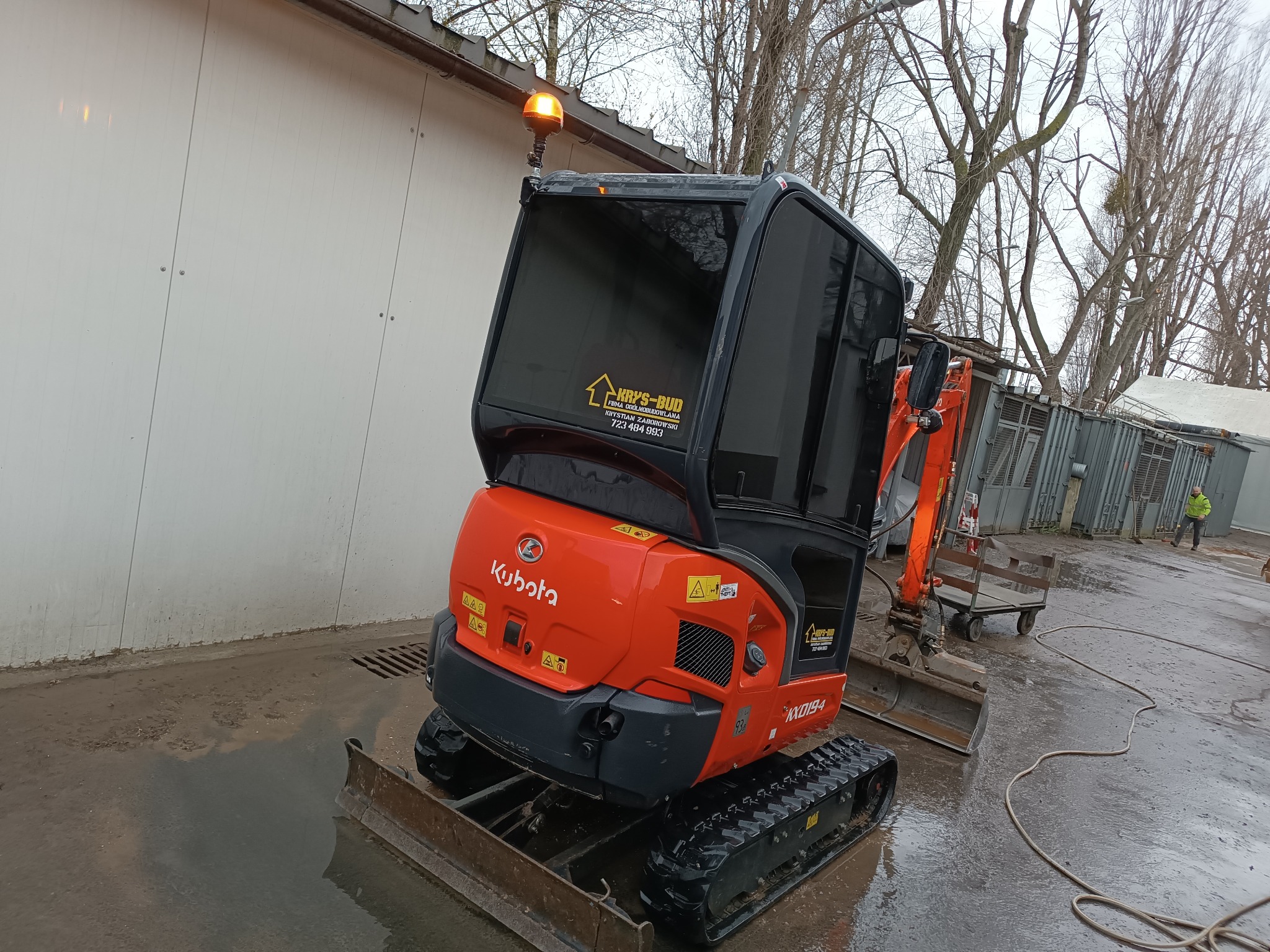 Pomarańczowa minikoparka Kubota KX019-4 z włączonym światłem ostrzegawczym na mokrym placu, z widocznym logo firmy Krys-Bud na kabinie i osobą w tle.