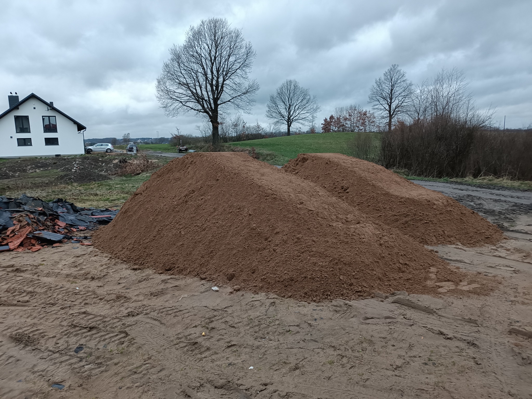 Dwie pryzmy brązowego materiału na ziemi, prawdopodobnie piasku lub ziemi, z widocznymi śladami opon, w tle dom z białymi ścianami i szarym dachem oraz drzewa bez liści pod pochmurnym niebem.