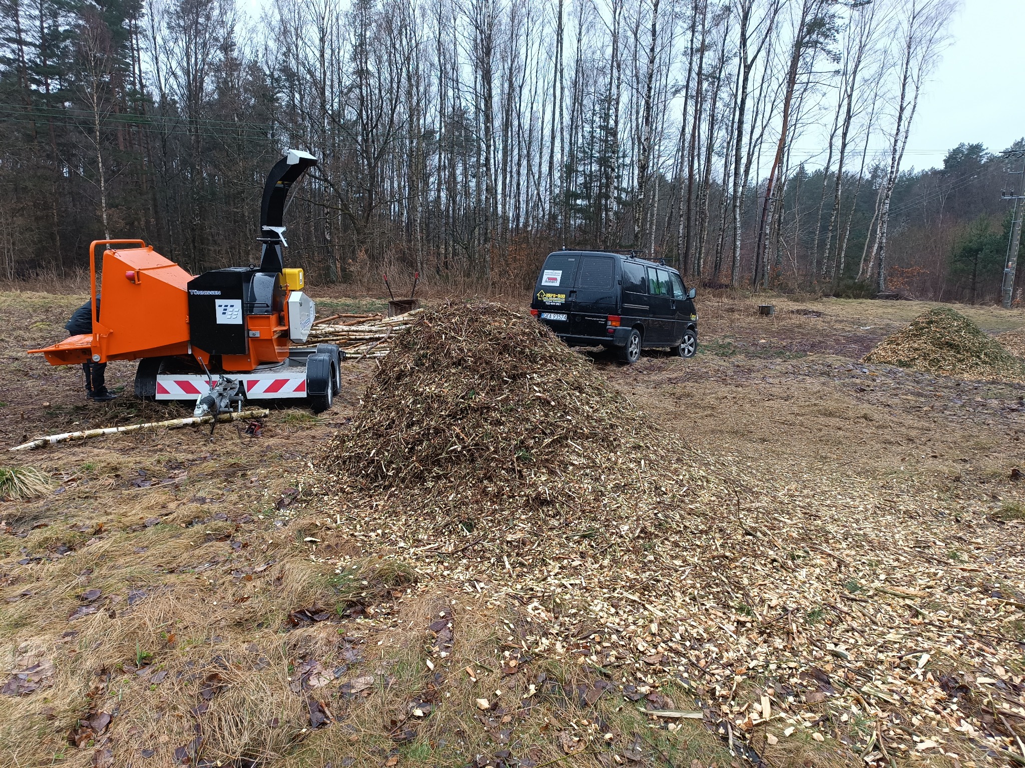 Pomarańczowa rębarka do gałęzi na przyczepie, obok stos świeżych zrębków i czarny bus firmy na skraju lasu w pochmurny dzień.