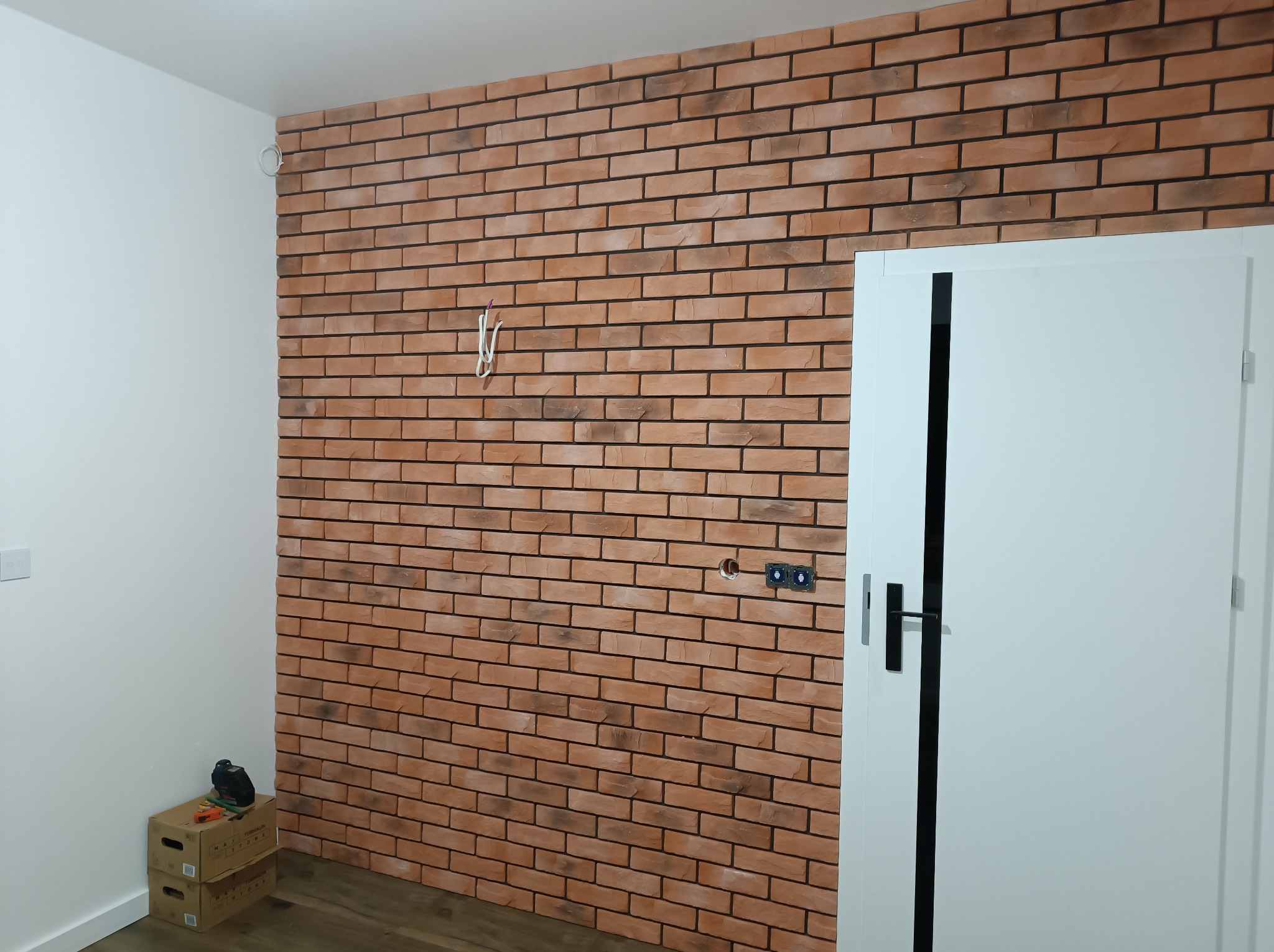 Ściana wewnętrzna wyłożona cegłą klinkierową w odcieniach czerwieni i brązu z czarną fugą, widoczne puszki elektryczne i wystające przewody, obok białe drzwi z czarną klamką, na podłodze panele,...